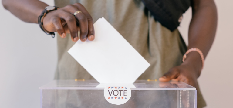Eine schwarze Person wirft ein Blatt Papier in eine Wahlurne. 